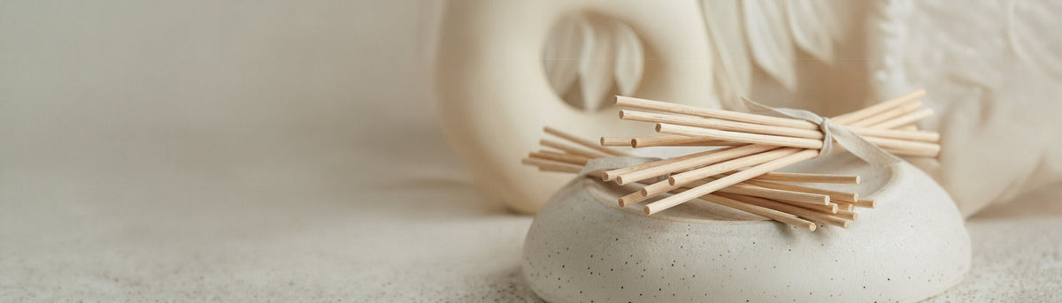 Ein mit einem Band verschnürtes Bündel von Schilfrohr-Diffusionsstäbchen aus Holz ruht auf einem glatten, runden Keramikhalter mit abstraktem Keramikdekor im Hintergrund auf einer neutralen Oberfläche.