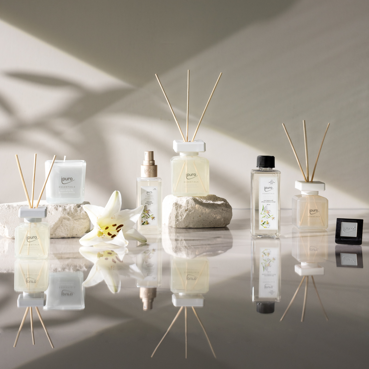Das ESSENTIALS ipuro white lily Raumspray von ipuro wird auf weißen Steinen mit Maiglöckchen und weißer Blüte präsentiert, die sich bei sanftem Tageslicht auf einer glänzenden Oberfläche spiegeln.