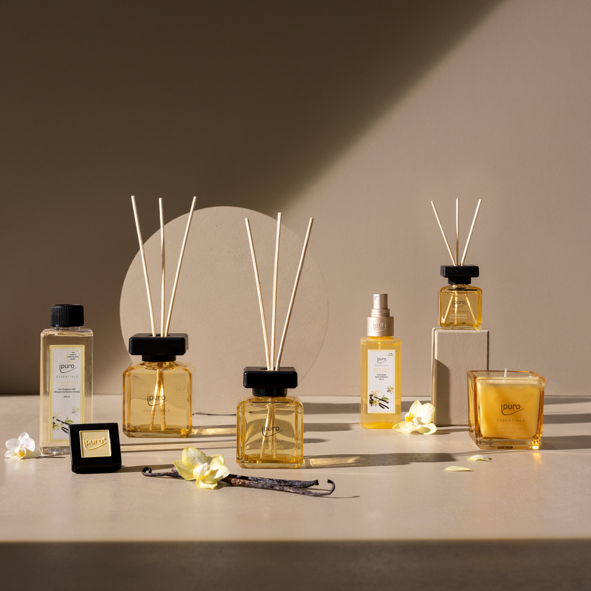 Auf einer beigen Fläche steht der ipuro ESSENTIALS soft vanilla Raumduft-Nachfüller von ipuro, ein bernsteinfarbener Raumduft mit Sandelholz und Vanille, neben Schilfrohrdiffusoren, einer Kerze, einem Spray, Dekostäbchen und weißen Blüten.