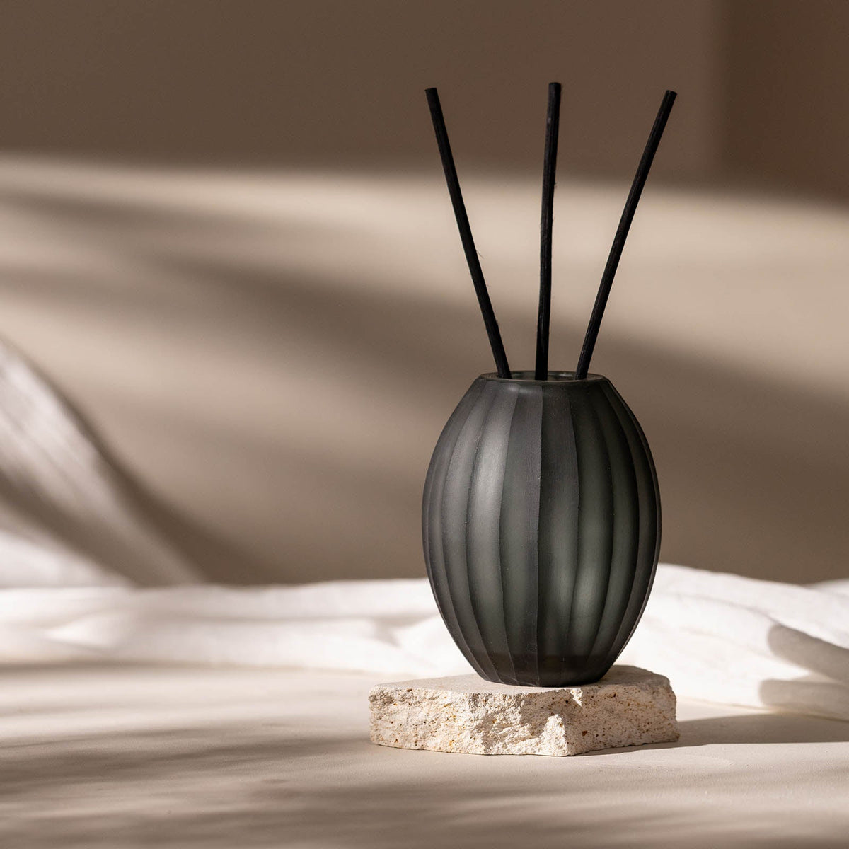 Der Atelier by ipuro gris Diffusor von ipuro mit einer gerippten Flasche aus dunklem Glas und drei schwarzen Stäbchen wird auf strukturiertem Stein vor einem sanft beleuchteten beigen Hintergrund mit natürlichem Licht und sanften Schatten präsentiert.