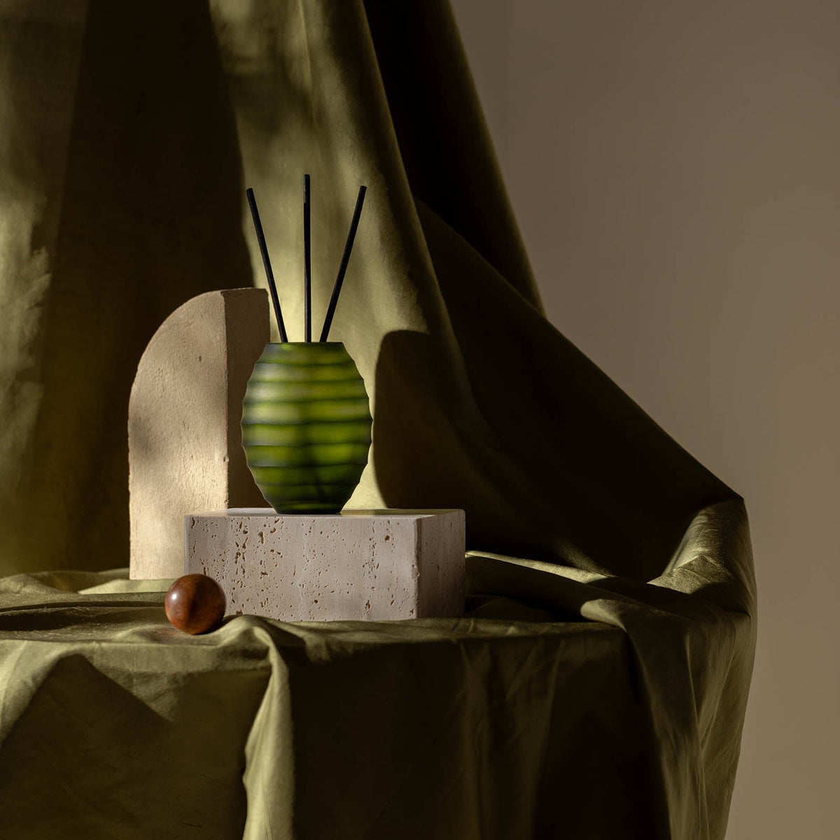 Ein Atelier by ipuro vert Diffusor mit schwarzem Stäbchen von ipuro ruht auf einem Steinblock neben einem geschwungenen beigen Objekt und einer braunen Kugel, die auf einem drapierten olivgrünen Stoff angeordnet sind und für auffällige Licht- und Schatteneffekte sorgen.