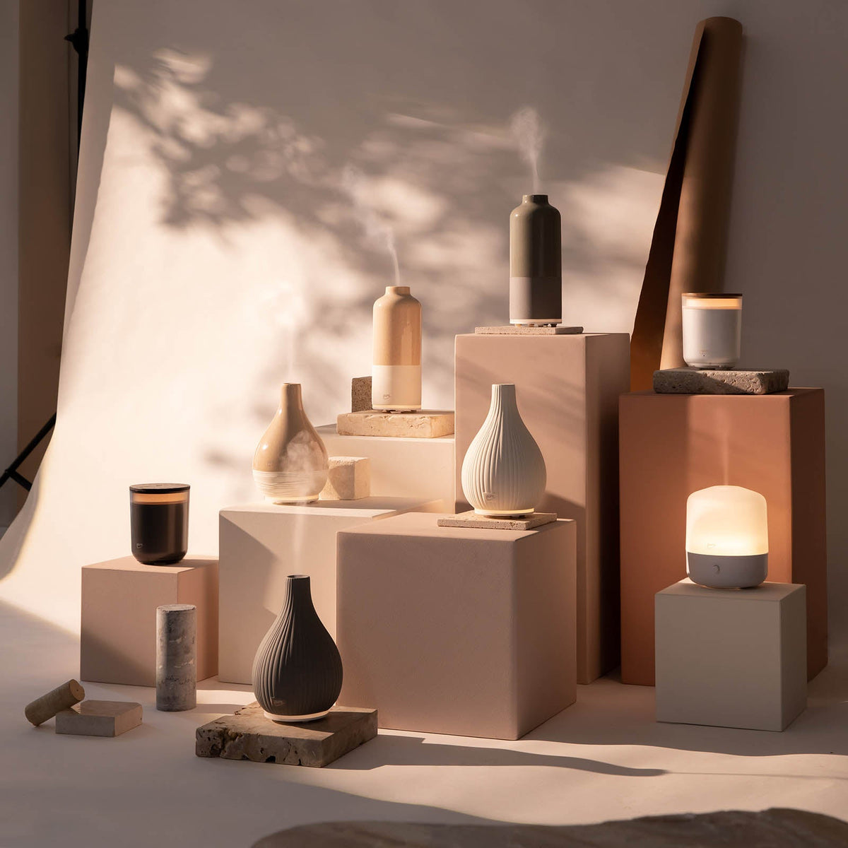 Die ipuro AIR SONIC Aroma-Vase Elektrischer Aroma-Diffusor und Kerzen in neutralen Farbtönen von ipuro stehen auf beigen und weißen Sockeln, die von sanftem Sonnenlicht beleuchtet werden und eine beruhigende Wirkung haben.