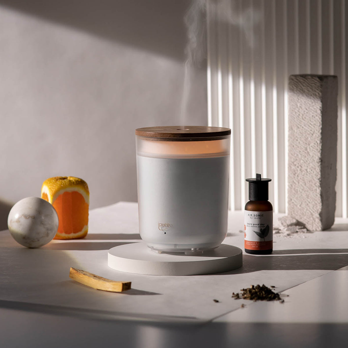 Die moderne weiße AIR SONIC ipuro Aromakerze Elektrischer Aroma-Diffusor von ipuro hat einen Holzdeckel und einen runden Sockel, der mit einer kleinen bernsteinfarbenen Tropfflasche, einer Orangenscheibe, einer Marmorkugel und strukturierten Steinen in sanftem Tageslicht gezeigt wird.