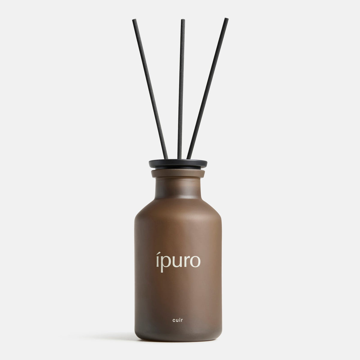 Ein brauner Milchglasflakon mit der Aufschrift "ipuro" (Raumduft ipuro cuir) und drei schwarzen Schilfrohren verströmt einen feinen Duft von Vanille und Zitrusnoten, der sich von einem schlichten weißen Hintergrund abhebt.