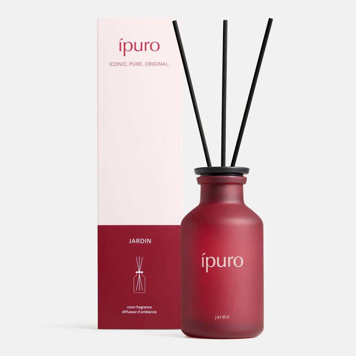 Ein mattroter Glasflakon mit der Aufschrift ipuro und drei schwarzen Schilfrohren steht neben einer passenden rosa-weißen Schachtel - beide aus dem ipuro Raumduft CLASSIC jardin, der den Duft exotischer Blüten einfängt und an einen verborgenen Garten erinnert.