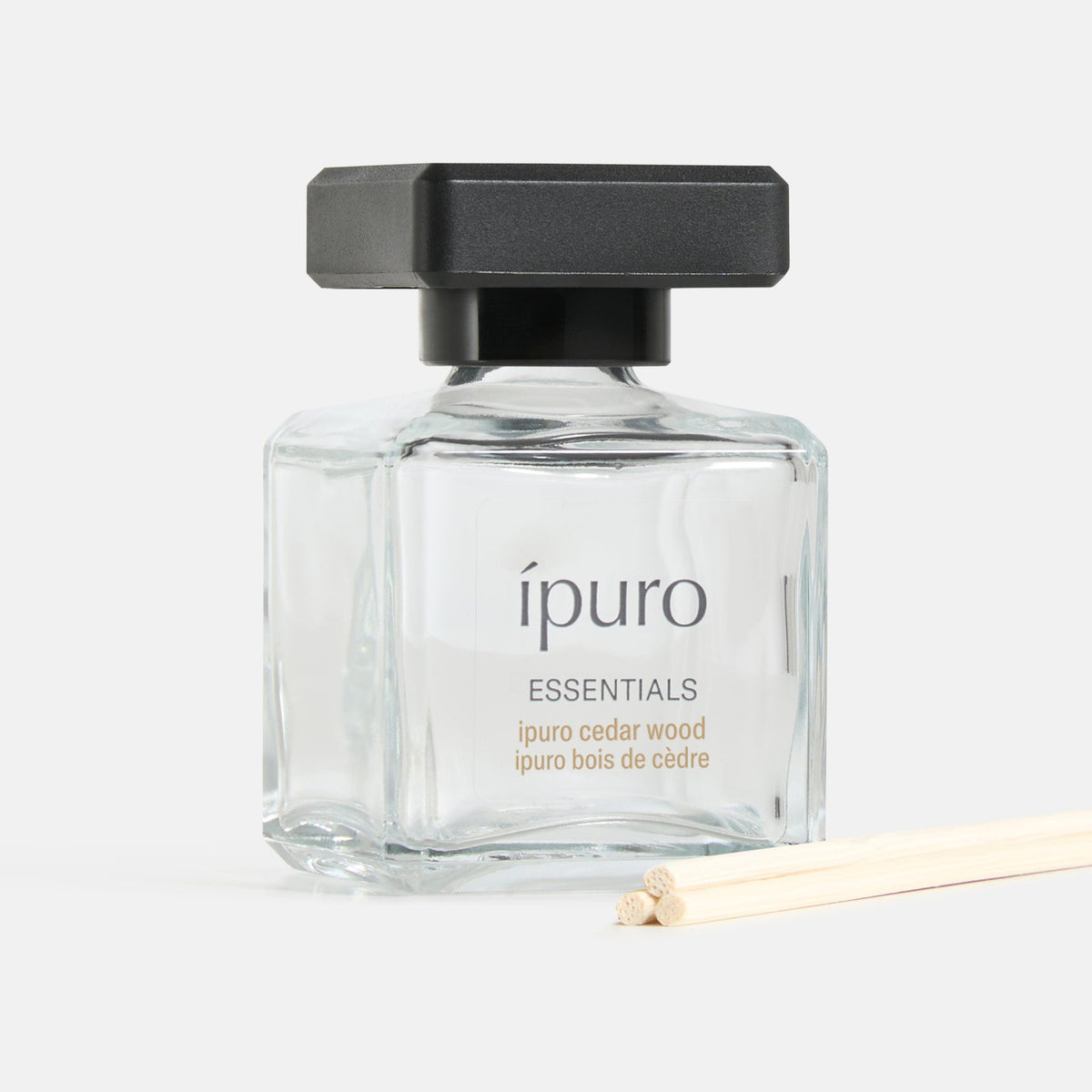 Der Raumduft ipuro cedar wood von ipuro präsentiert sich in einem klaren Glasflakon mit schwarzer, rechteckiger Kappe, begleitet von dünnen Holzstäbchen auf weißem Grund, die einen holzigen Duft mit subtilen blumigen Noten verströmen.