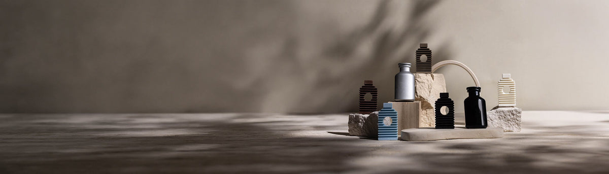 Minimalistisches Display mit verschiedenen Flaschen und gestreiften Behältern, die auf strukturierten Steinblöcken angeordnet sind, mit weichen, natürlichen Schatten vor einem neutralen Hintergrund.