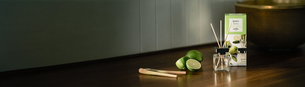 Ein Schilfrohrdiffusor, seine Box, frische Limetten und Zitronengras stehen auf einer Holzfläche neben einer großen Messingschale und schaffen eine ruhige, natürliche Atmosphäre.