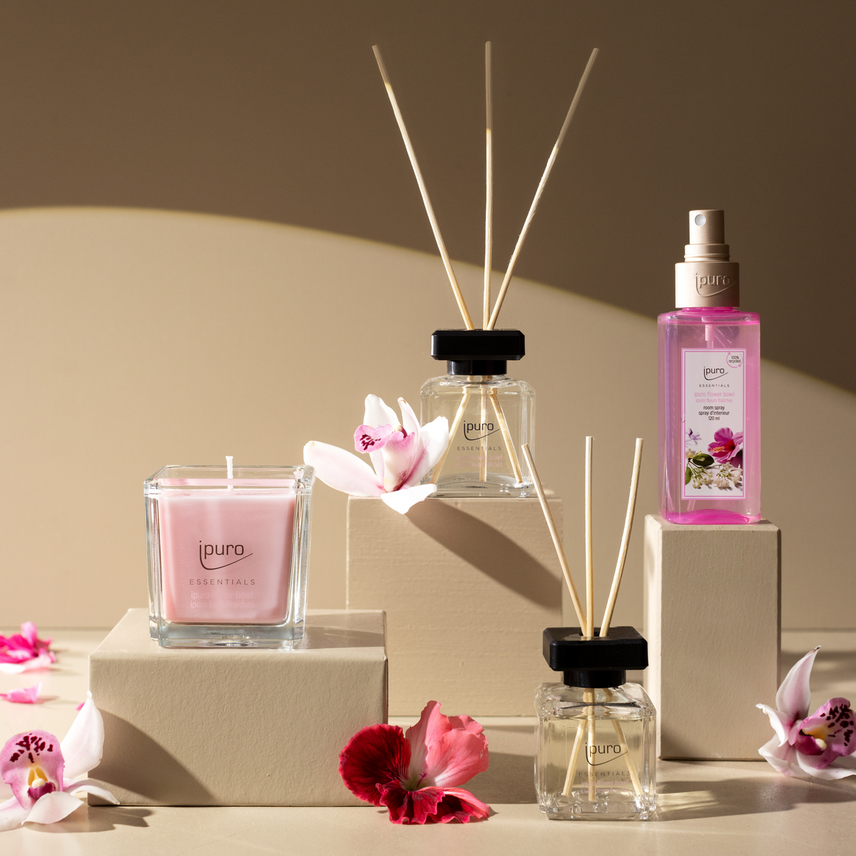 Eine Auslage von ipuro-Duftprodukten - einschließlich der Autoduft ipuro-Blumenschale - auf beigen Blöcken mit rosa Blumen schafft eine blumige Note und belebende Frische vor neutralem Hintergrund.