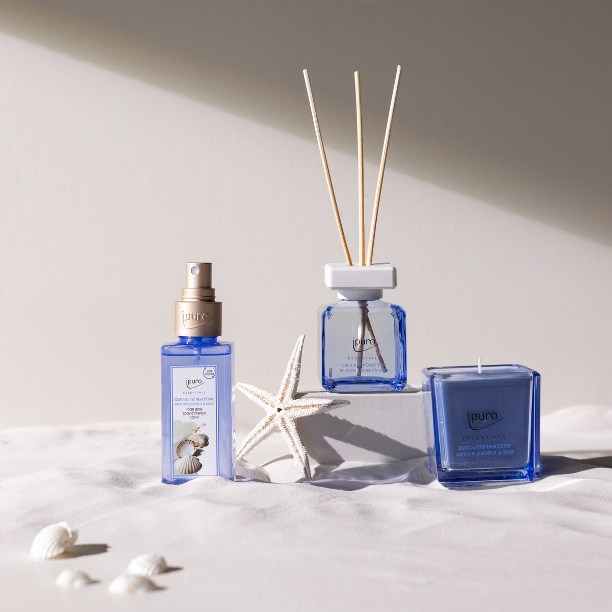 Die ipuro Duftkerze "sunny beachtime" von ipuro liegt auf weißem Sand mit Muscheln und einem Seestern und fängt Sommer am Strand-Stimmung unter sanftem Sonnenlicht vor einem blassbeigen Hintergrund ein.