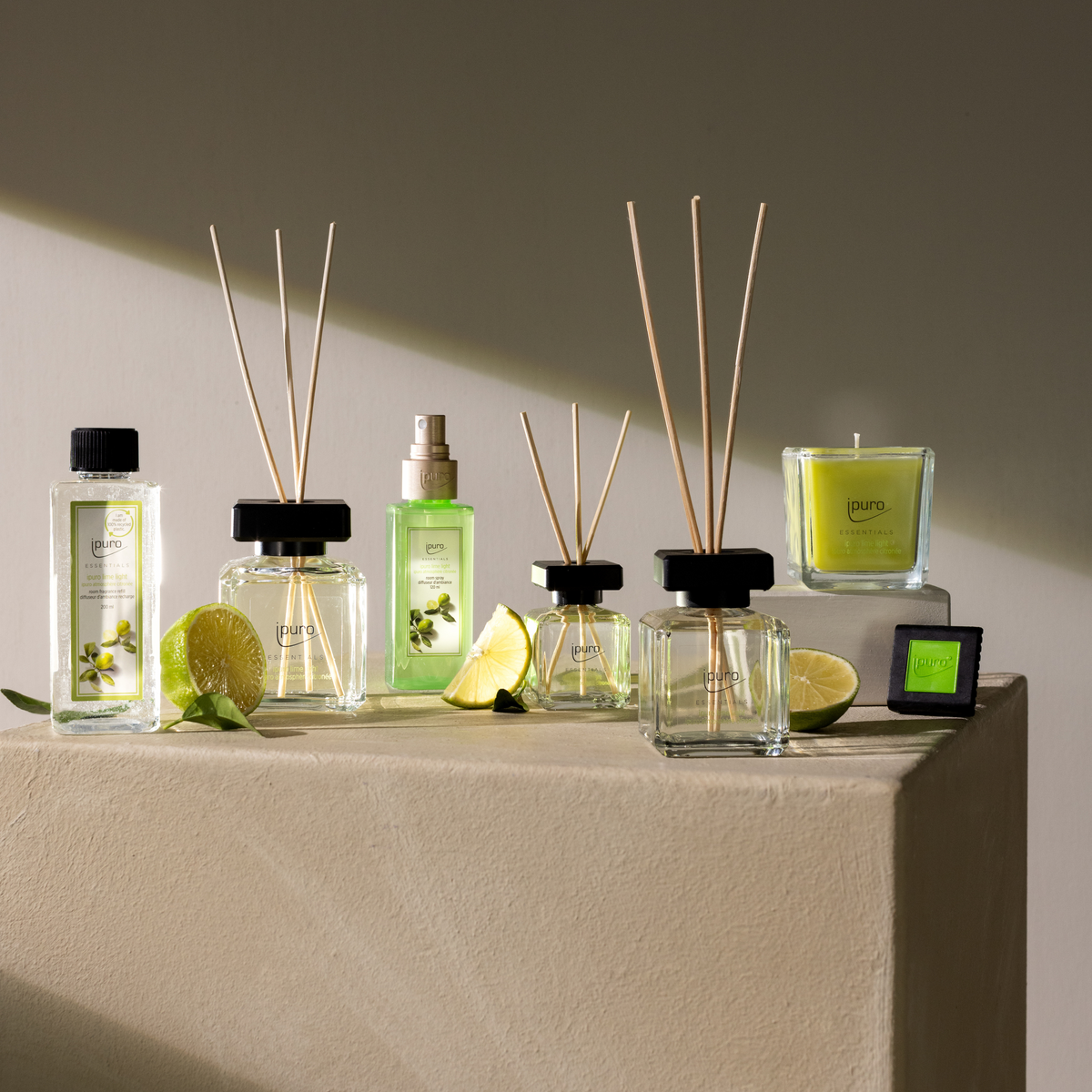 Experience the ESSENTIALS ipuro lime light Raumspray, showcased with reed diffusers, a candle, and sliced limes on a beige pedestal, all in soft natural light for a fresh green-themed home ambiance.