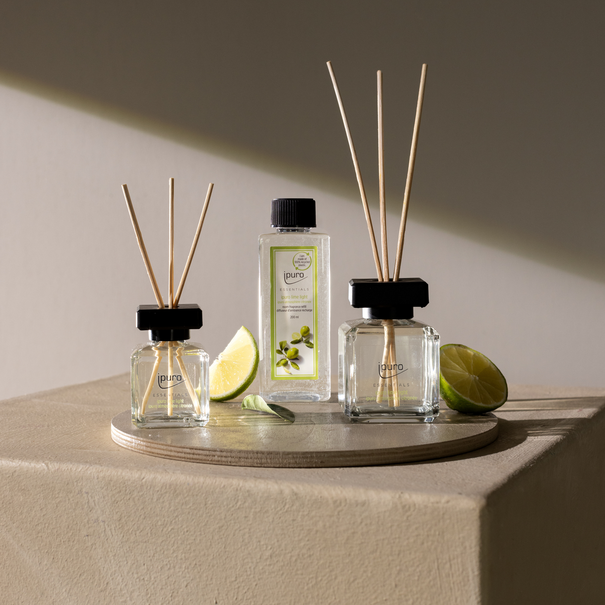 The ESSENTIALS ipuro lime light Raumspray is shown on a round tray beside glass reed diffusers, lime wedges, and slices, all arranged on a beige surface in natural light.