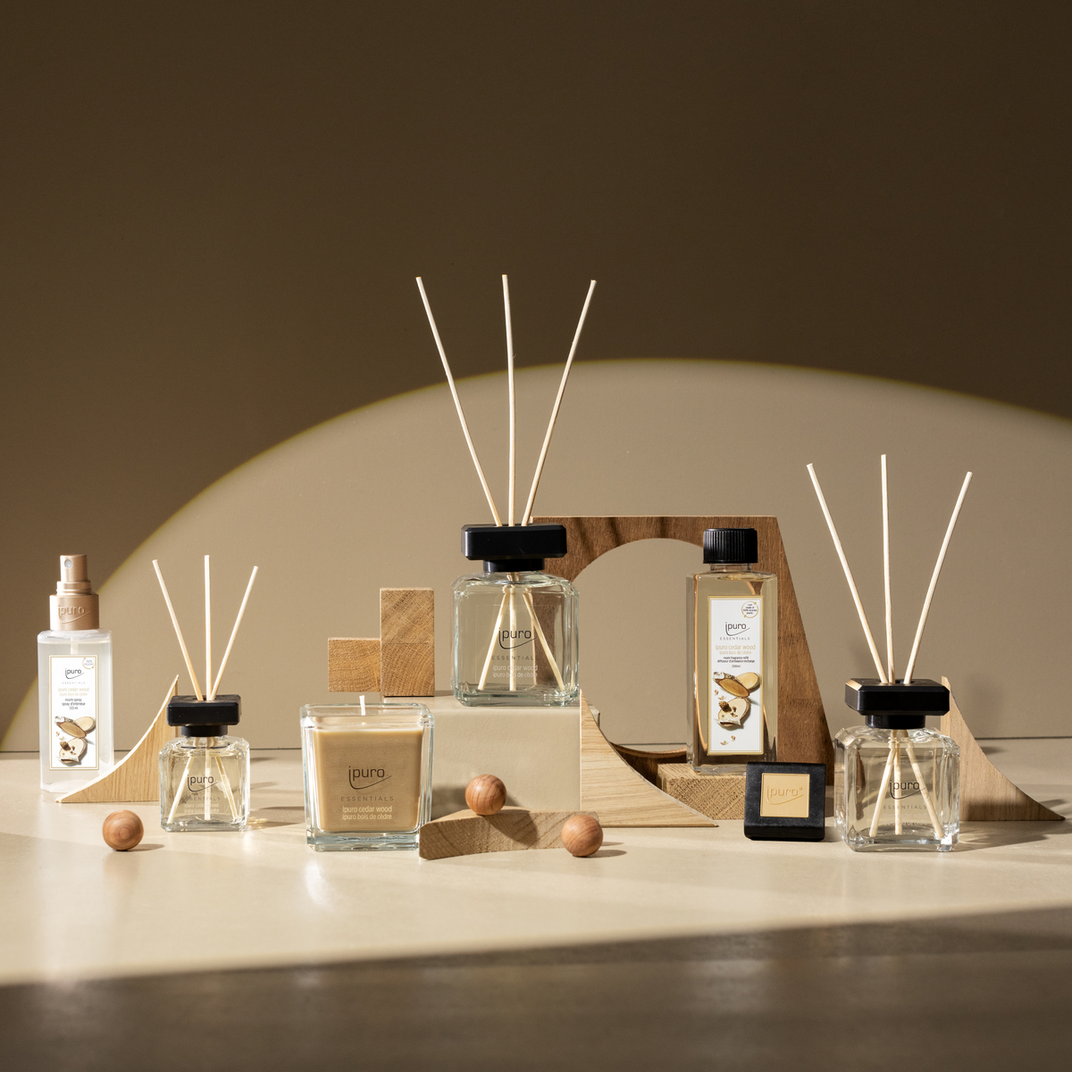 The ESSENTIALS ipuro cedar wood Raumspray, along with other home fragrance items featuring woody notes, is displayed on geometric wooden shapes with decorative wooden balls and illuminated by soft natural light.