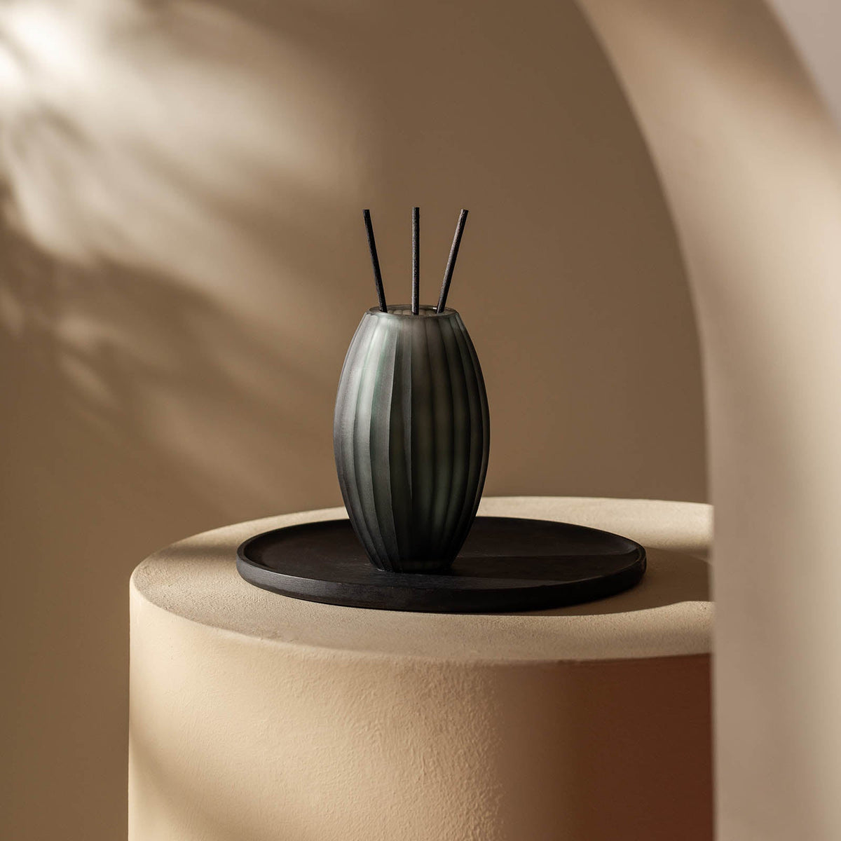 Der gerippte, dunkle Glasdiffusor Atelier by ipuro gris von ipuro mit drei schwarzen Schilfrohren steht auf einem runden Tablett auf einem cremefarbenen Sockel und wird sanft beleuchtet - eine Szene, die die Eleganz der Atelier by ipuro Design-Vasen widerspiegelt.