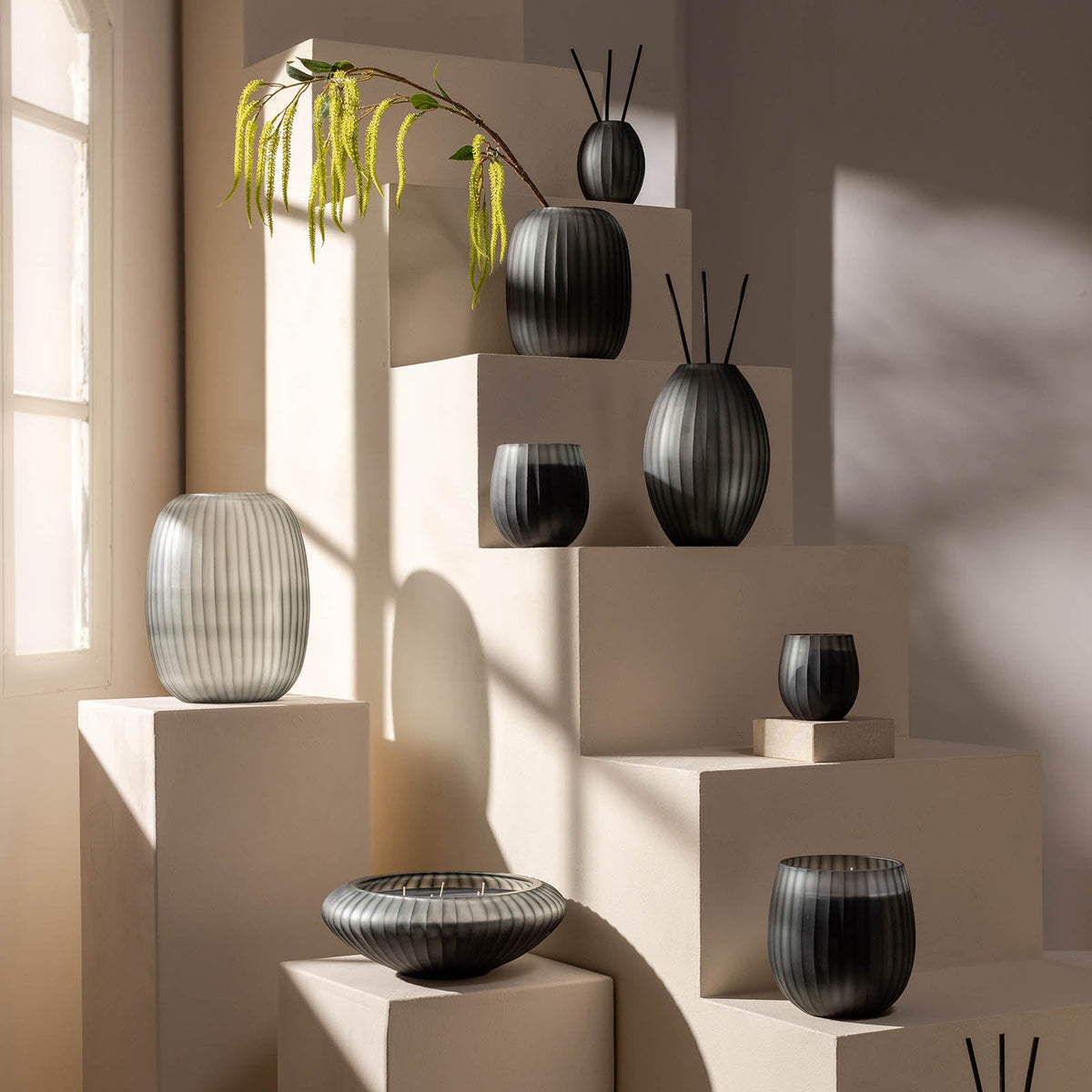 Der Atelier by ipuro gris Diffusor von ipuro mit seinem gerippten rauchgrauen Design steht stilvoll auf einem cremefarbenen Sockel. Das einfallende Sonnenlicht und ein Zweig mit grünen Blättern vervollständigen den modernen Look - werten Sie Ihren Raum mit diesem eleganten Duftobjekt auf.