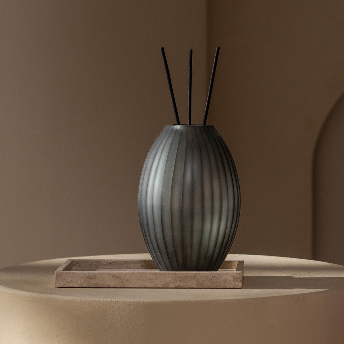 Ein dunkler, gerippter Atelier by ipuro gris Diffusor mit drei schwarzen Stäben sitzt auf einem rechteckigen Steintablett auf einer runden, beigen Fläche, sanft beleuchtet vor einem minimalistischen Hintergrund - ein raffinierter Akzent von ipuro.