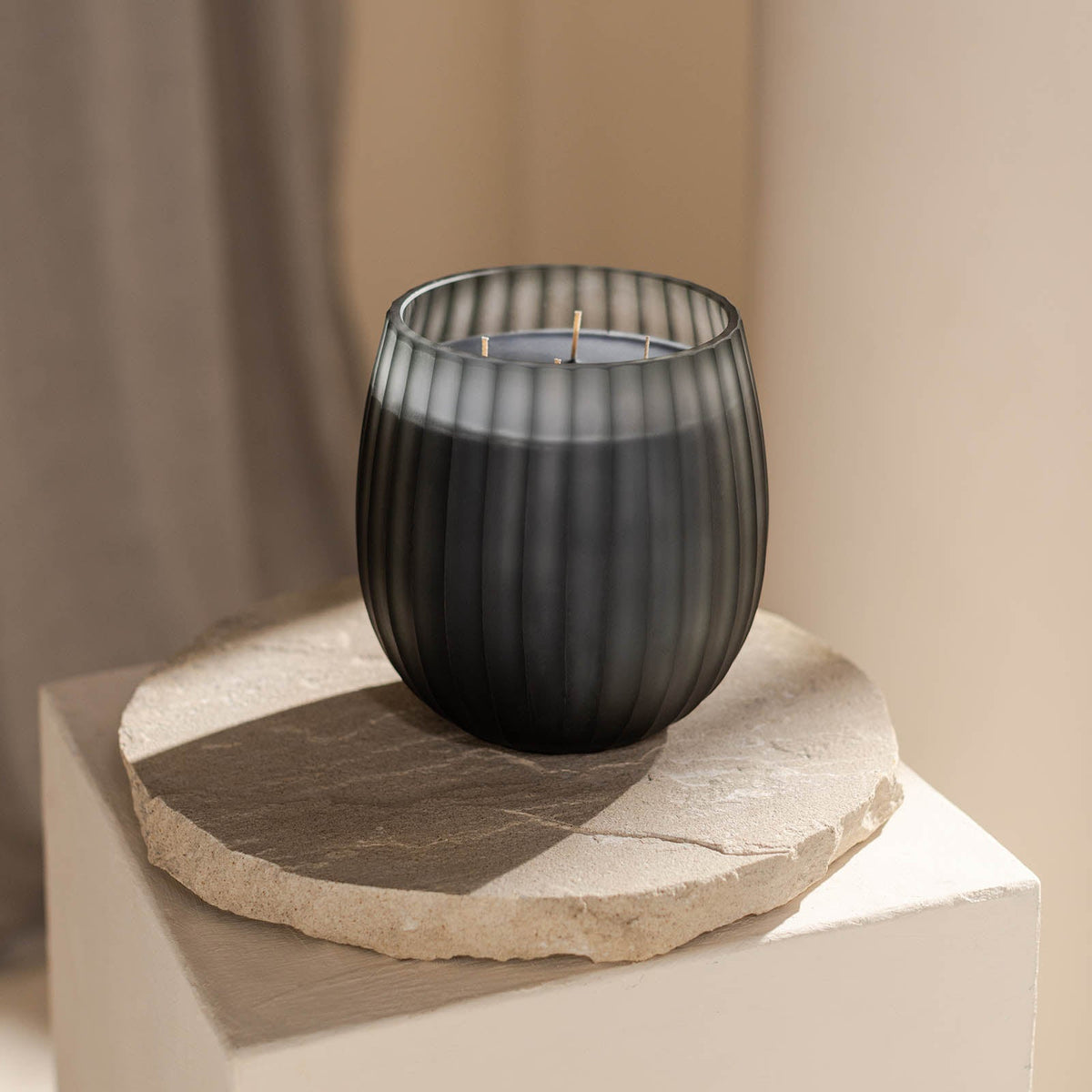 Die Atelier by ipuro gris Duftkerze von ipuro, eine schwarze gerippte Glaskerze mit drei Dochten, ruht auf einer runden Steinplatte auf einem leichten Sockel - perfekt für Fans von luxuriösen Kerzen und minimalistischem Design.