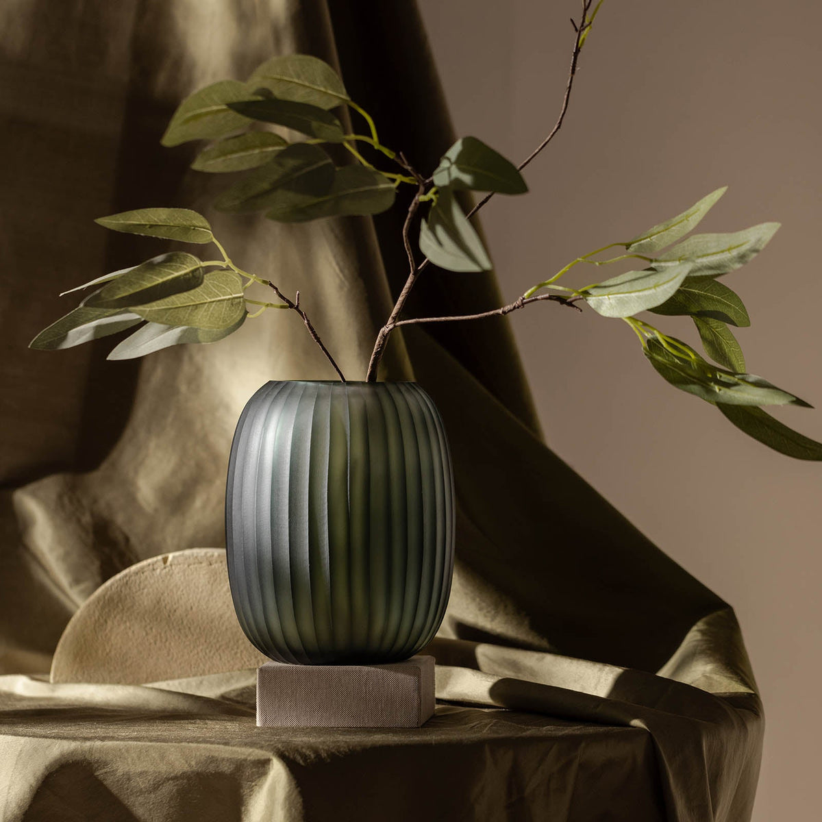 Die Vase Atelier by ipuro gris von ipuro zeigt ein grünes, geripptes Design mit blättrigen Zweigen auf einem quadratischen Steinsockel, gestylt auf seidig drapiertem Stoff in weichem, stimmungsvollem Licht für eine exklusive Designnote.