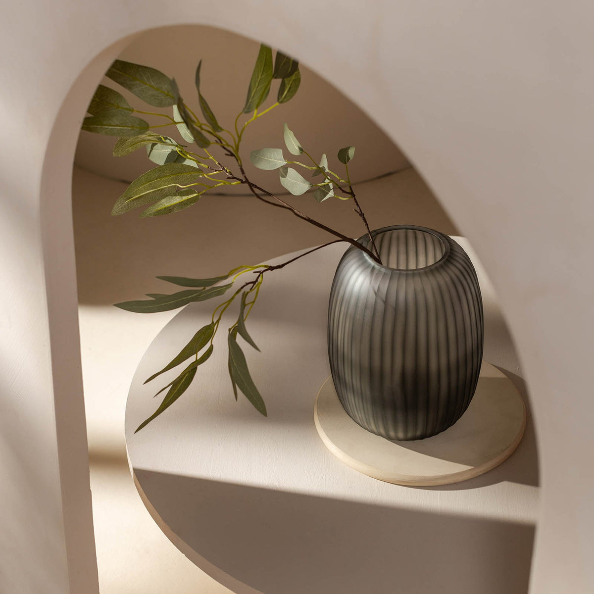 Die Vase Atelier by ipuro gris von ipuro mit ihrem gerippten grauen Design hält grüne, blättrige Zweige auf einer runden weißen Ablage unter einem geschwungenen Bogen, der in sanftem natürlichem Licht erstrahlt und einen exklusiven Stil präsentiert.