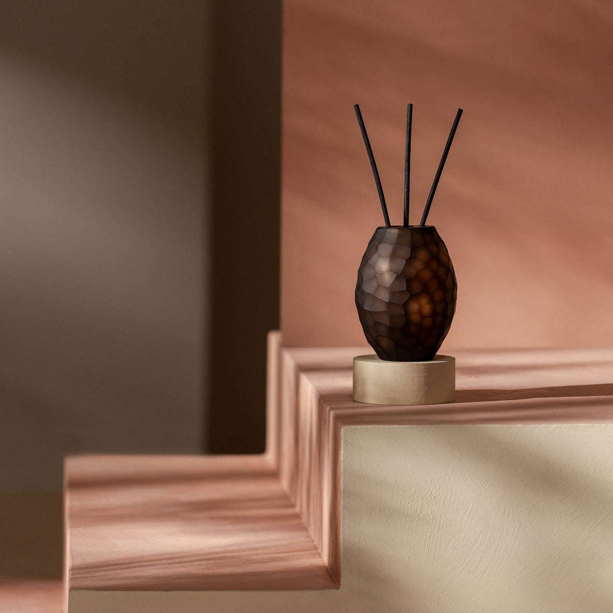 Der Atelier by ipuro brun Diffusor von ipuro mit seinem dunklen, strukturierten Design auf einem runden, beigen Sockel wertet Ihren Raum mit exklusiven Düften und einem raffinierten Stil auf, der sich in sanftem, natürlichem Licht auf blassblauen, geometrischen Stufen entfaltet.