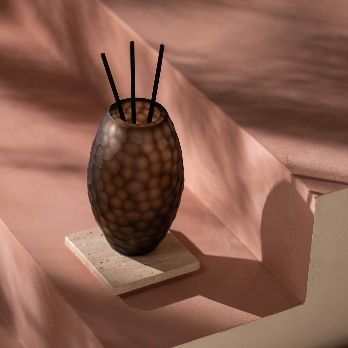 Der Atelier by ipuro brun Diffusor von ipuro mit einem strukturierten braunen Glasdesign mit schwarzen Stäben ruht auf einem hellen Steinuntersetzer auf einer rosa-beigen Stufenfläche in sanften Schatten.