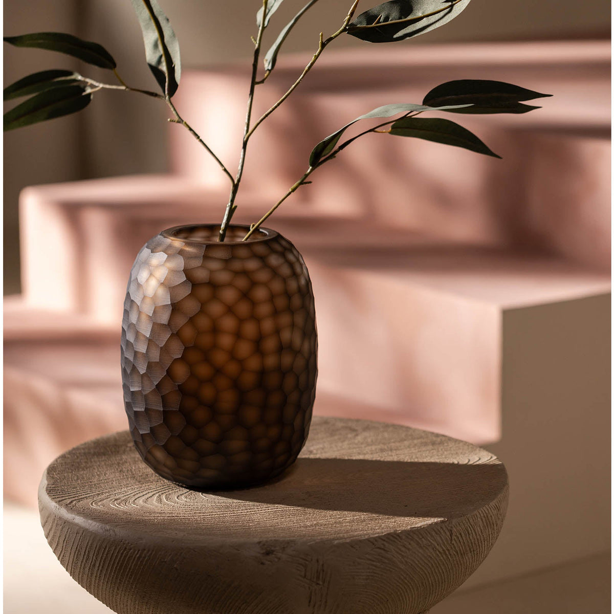 Die Vase Atelier by ipuro brun von ipuro mit Wabenstruktur hält einen belaubten Zweig auf einem runden Holztisch. Sanftes Sonnenlicht wirft Schatten auf die blassrosa Stufen im Hintergrund.