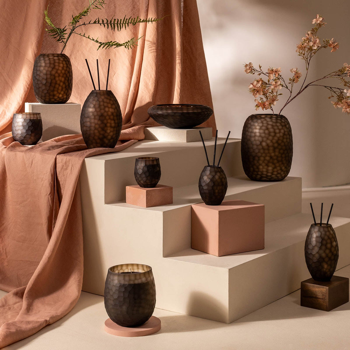 Eine Ausstellung von Atelier by ipuro brun Vasen und Diffusoren von ipuro steht auf cremefarbenen Stufen, hinter denen brauner und pfirsichfarbener Stoff drapiert ist; einige Vasen sind mit zarten grünen und rosa Zweigen versehen.