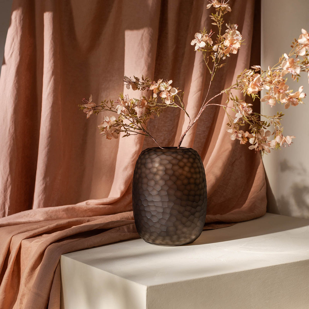 Die ipuro Atelier by ipuro brun Vase zeigt ein strukturiertes, rauchiges Design mit zarten rosafarbenen Kirschblütenzweigen auf einer weißen Oberfläche mit zartem pfirsichfarbenem Stoff - ein stilvolles Stück, das sich ideal für elegante Innenräume eignet.