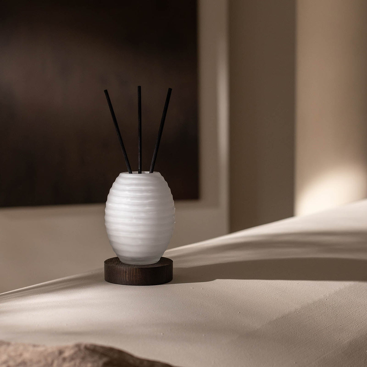 Der Atelier by ipuro blanc Diffusor von ipuro, eine weiße gerippte Keramik-Duftvase, ruht auf einem runden Holzsockel vor einem sanft beleuchteten, neutralen Hintergrund, der die Eleganz der exklusiven Duftvasen unterstreicht.