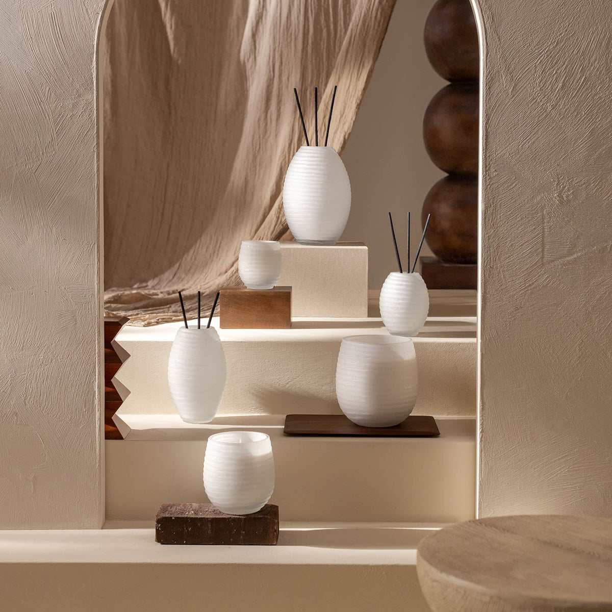 Ein minimalistisches Arrangement aus weißen, gerippten Keramikvasen und der Atelier by ipuro blanc Duftkerze von ipuro steht auf gestuften beigen Podesten vor strukturierten Wänden und neutral drapierten Stoffen und schafft eine ruhige und elegante Atmosphäre.