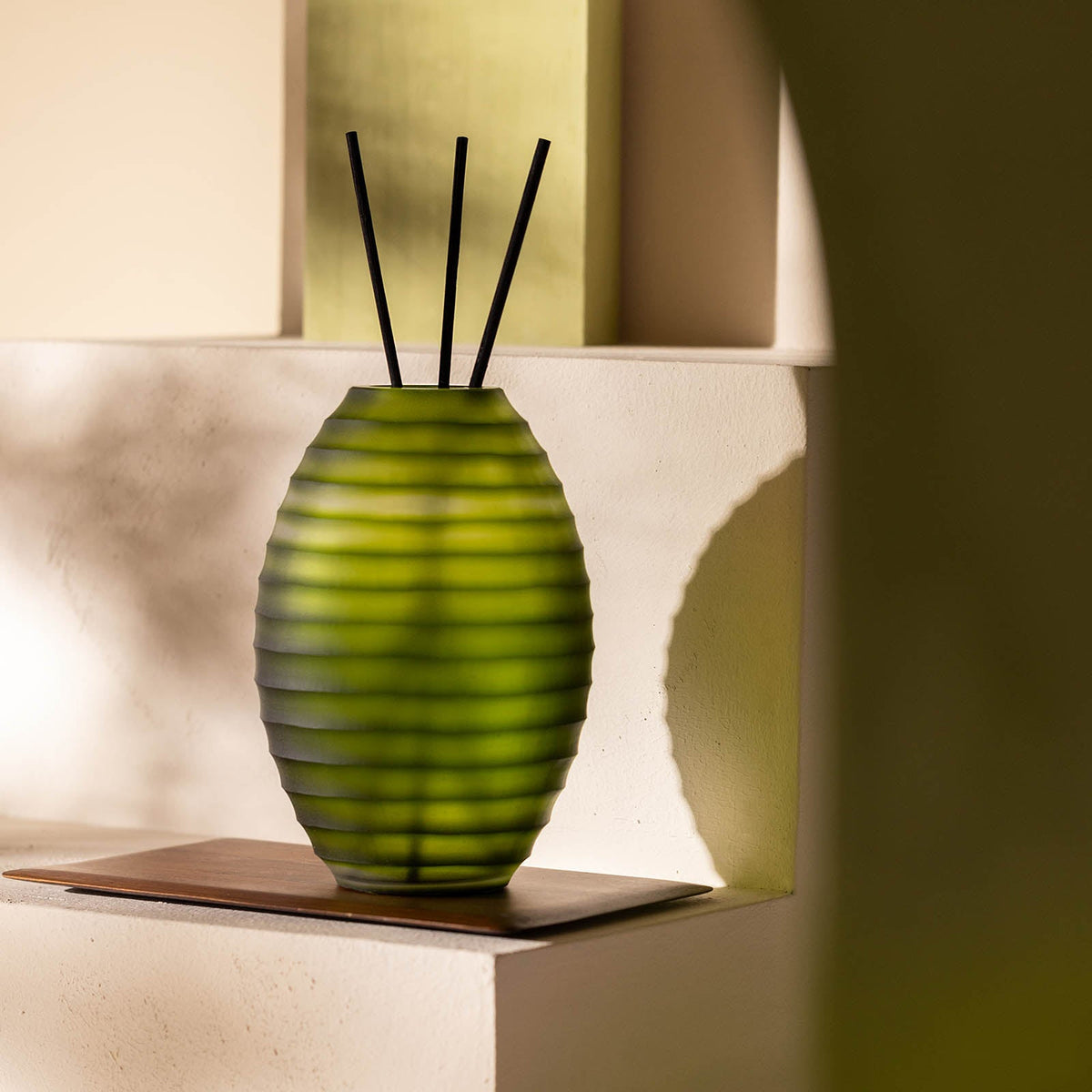Der Atelier by ipuro vert Diffusor von ipuro hat ein grünes, geripptes Glasdesign mit drei schwarzen Schilfrohren, die auf einem Holztablett auf einer weißen Oberfläche präsentiert werden - sein eleganter Stil ist ideal für die Präsentation luxuriöser Düfte.