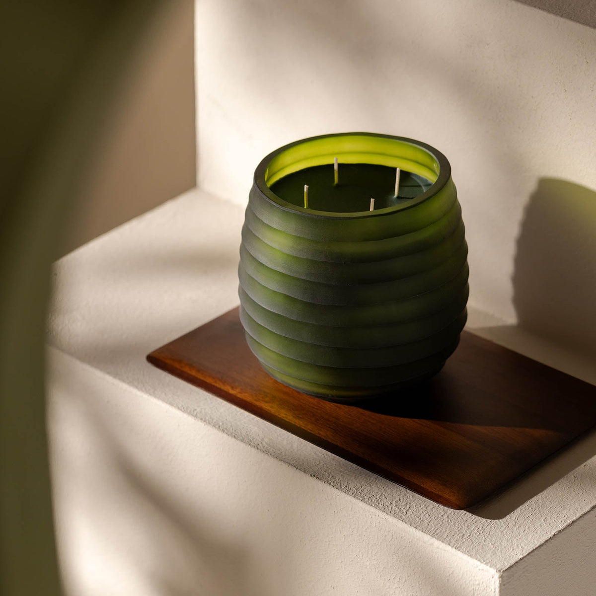 Die Atelier by ipuro vert Duftkerze von ipuro, eine grüne gerippte Glaskerze mit mehreren Dochten, ruht auf einem Holztablett auf einer leicht gestuften, in natürliches Licht getauchten Fläche, ideal für Fans von handgefertigten Kerzen.