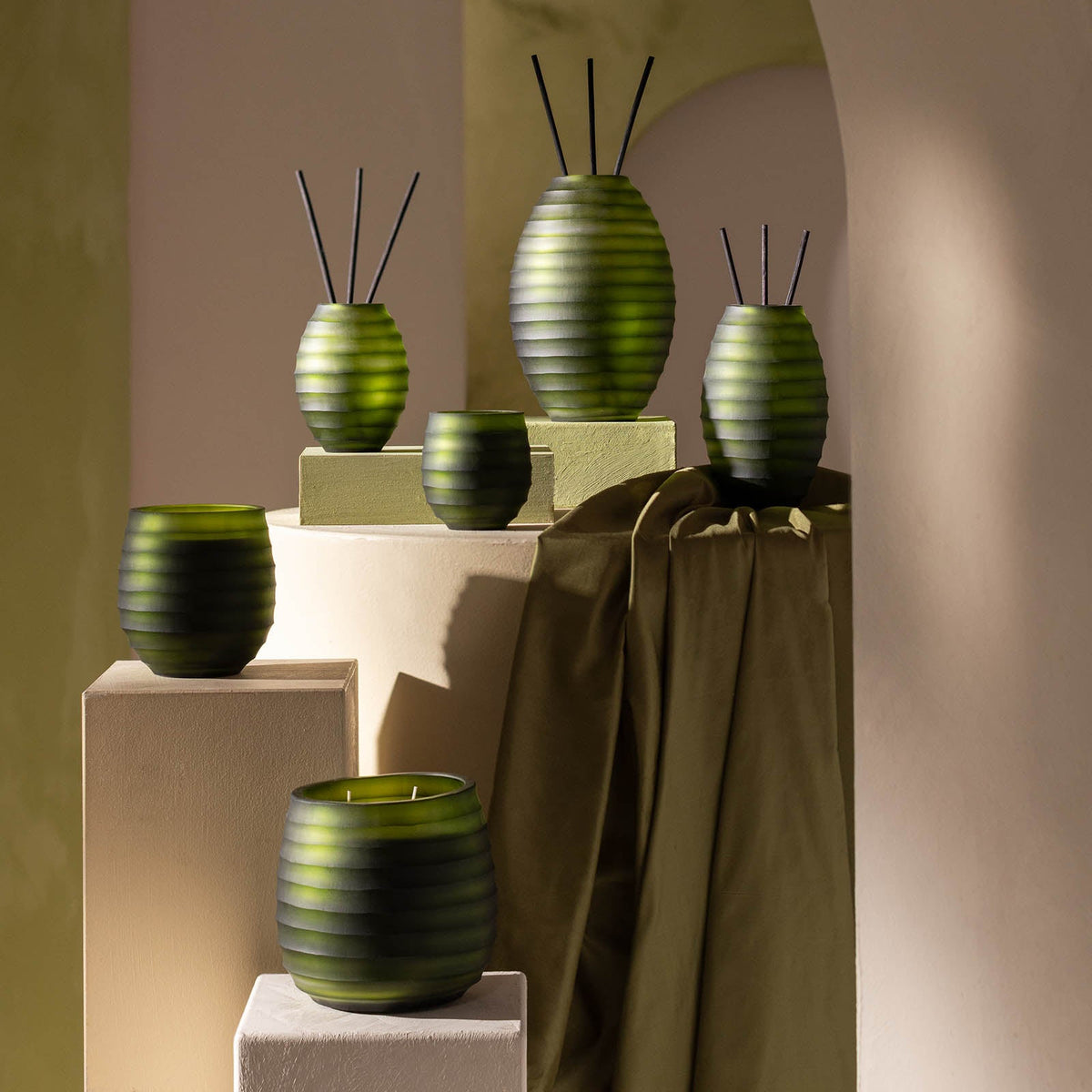 Ein Display mit Atelier by ipuro vert Raumduft von ipuro, mit grünen gerippten Glasbehältern in verschiedenen Größen mit schwarzen Schilfrohrstäben, auf beigen Ständern und olivgrünem Stoff im Hintergrund.
