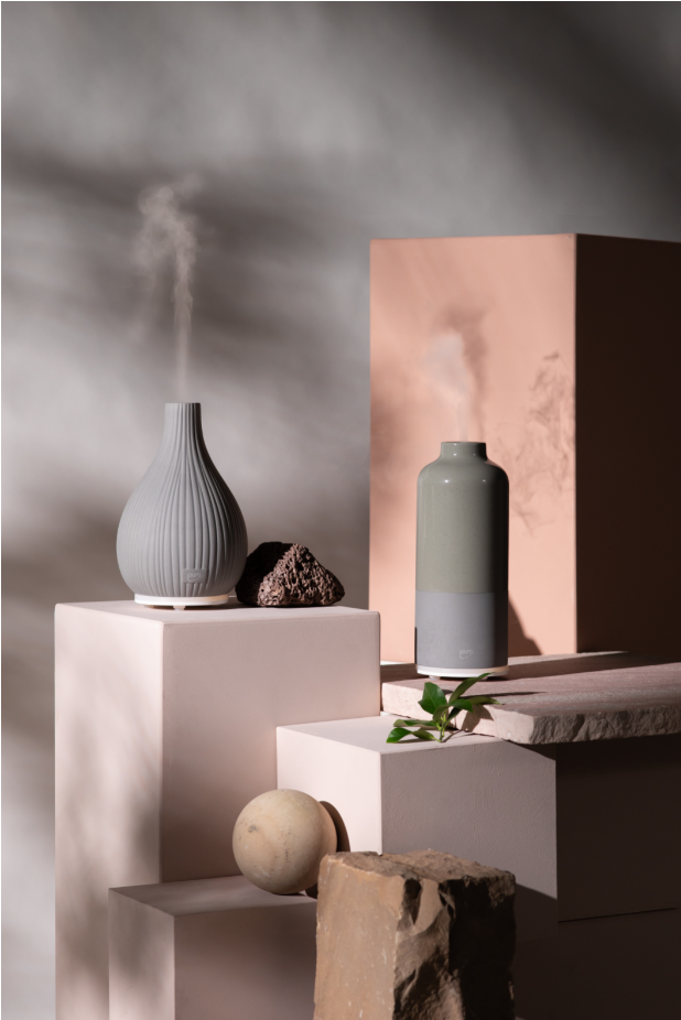 Die AIR SONIC ipuro Aroma-Flasche Elektrischer Aroma-Diffusor von ipuro verströmt Nebel auf einem hellrosa Sockel, umgeben von Steinen, grünen Blättern und einem runden Stein. Dramatische Schatten und natürliches Licht schaffen einen beruhigenden Raum für Ihre Lieblingsdüfte.