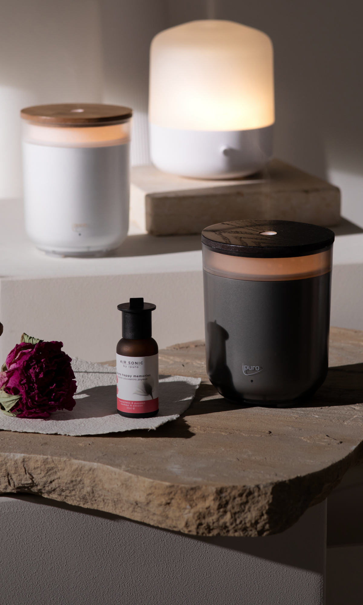 Ein schwarzer ipuro AIR SONIC Aroma-Stimmungsdiffusor, ein Duftöle-Fläschchen und eine getrocknete Rose ruhen auf steinernen und weißen Flächen, während zwei sanft leuchtende ipuro AIR SONIC Diffusoren im Hintergrund ein warmes, ruhiges Ambiente schaffen.