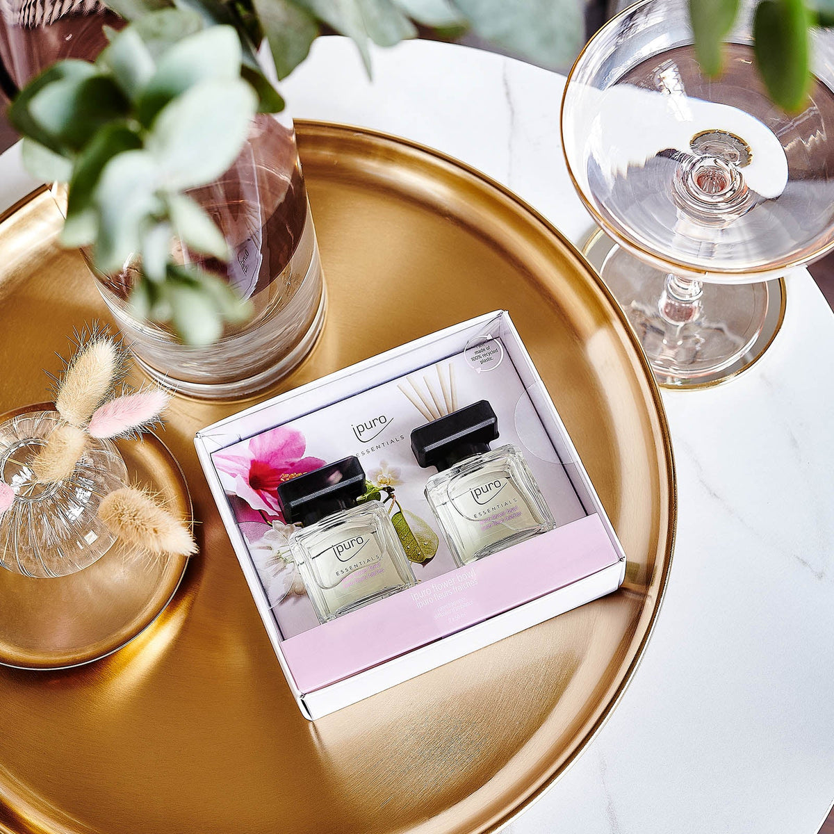 Auf einem goldenen Tablett wird das ipuro Raumduft-Set Blumenschale von ipuro präsentiert. Zwei Parfümflaschen mit schwarzem Deckel und hellem Etikett sowie eine Vase mit grünen Blättern und ein Glas sorgen für einen eleganten Look und einen einladenden Duft.