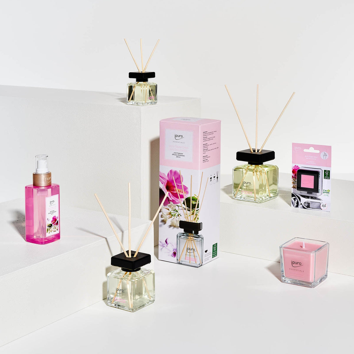 Das ESSENTIALS ipuro flower bowl Raumduft-Set von ipuro besteht aus einer blumigen Duftkerze, Spray und verpackten Gegenständen, die auf weißen Podesten arrangiert sind. In der Mitte befindet sich eine floral gestaltete Box für eine edle Blumennote.