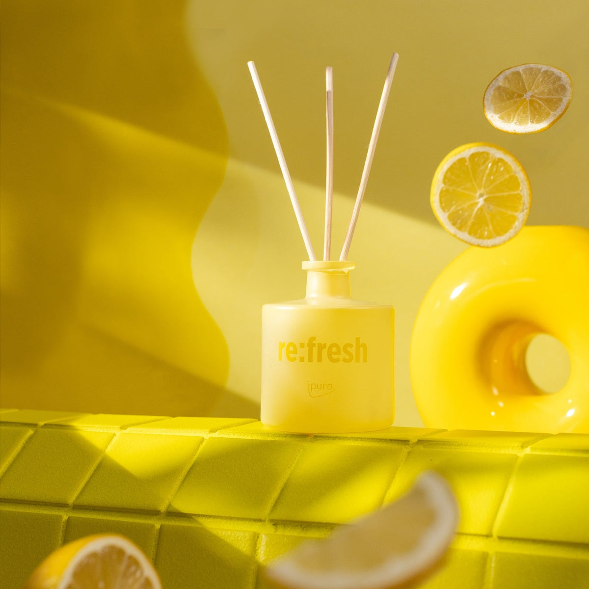 Der WE ARE: ipuro re:fresh Raumduft von ipuro, ein gelber Diffusor mit drei Stäben, steht auf gelben Fliesen inmitten von upgecycelten Zitronenscheiben und einem gelben Donut und schafft nachhaltige Frische vor einem leuchtend gelben Hintergrund.