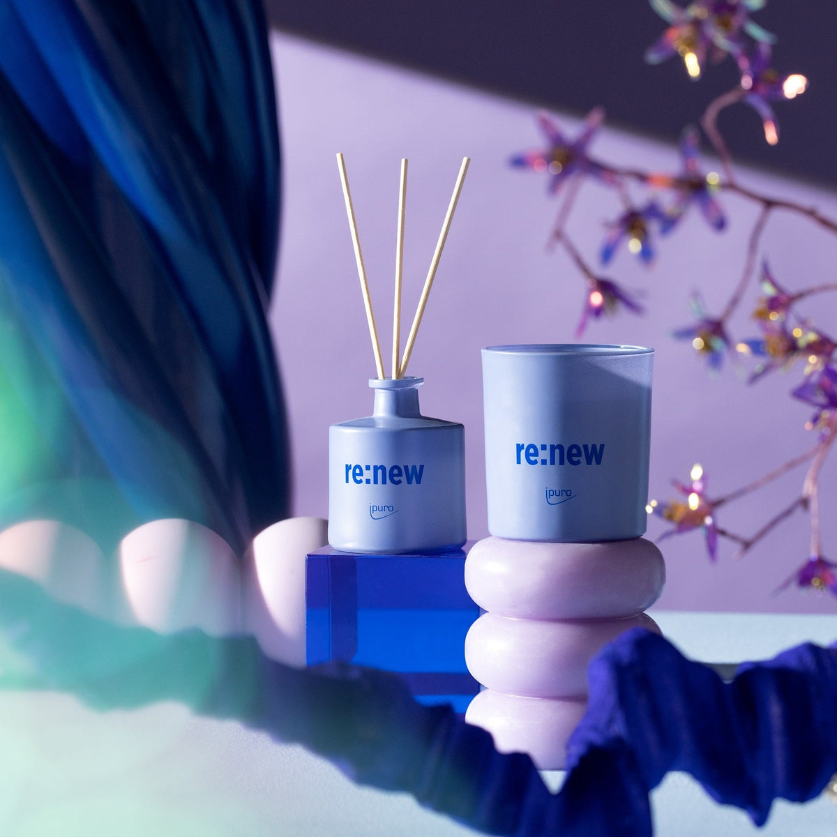 Der WE ARE: ipuro re:new Raumduft von ipuro mit Schilfrohrdiffusor und Kerze wird auf pastellrosa und blauen Podesten mit abstraktem Dekor und geretteten Blumen im Hintergrund bei sanfter, farbiger Beleuchtung gezeigt.