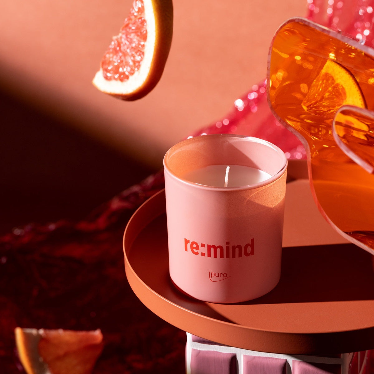 Die WE ARE: ipuro re:mind Duftkerze von ipuro in Pink mit Grapefruitduft wird auf einer runden Terrakottafläche zusammen mit Grapefruitscheiben und einem Glas Orangenflüssigkeit in einer warm beleuchteten Umgebung präsentiert.