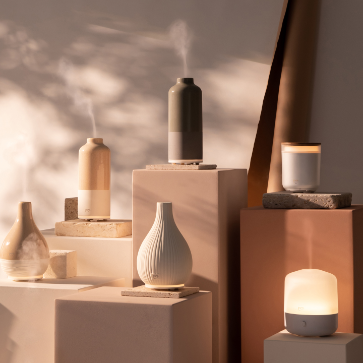 Ästhetische Präsentation von Keramik- und Glasdiffusoren, die Nebel ausstoßen, angeordnet auf Stein- und Sockelblöcken, mit weichem natürlichem Licht und Schatten, die eine beruhigende, minimalistische Atmosphäre schaffen.