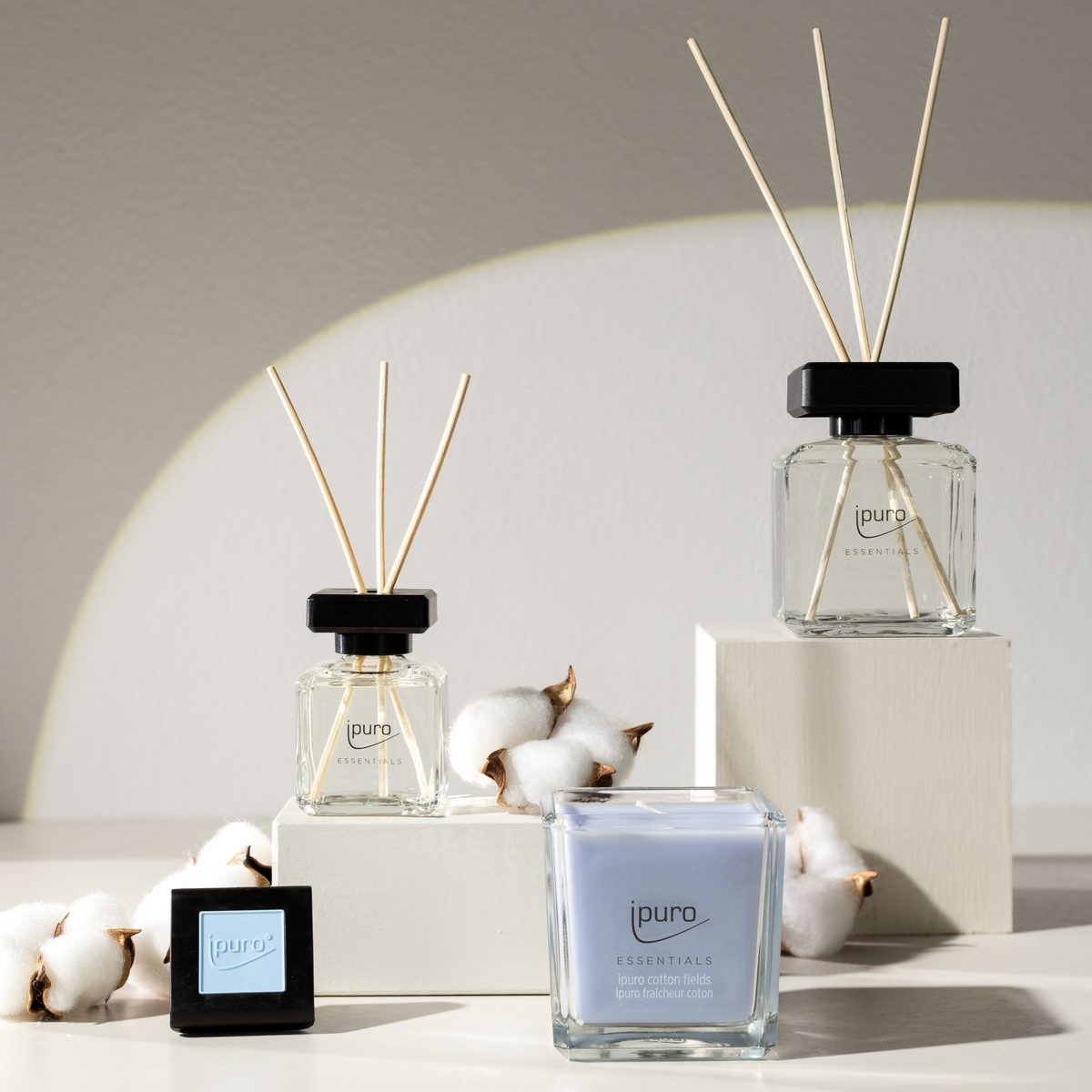 Der ESSENTIALS ipuro cotton fields Scent Plug Refill von ipuro ist auf weißen Blöcken mit Baumwollzweigen in einer hellen, minimalistischen Umgebung mit weichen Schatten an der Wand abgebildet.
