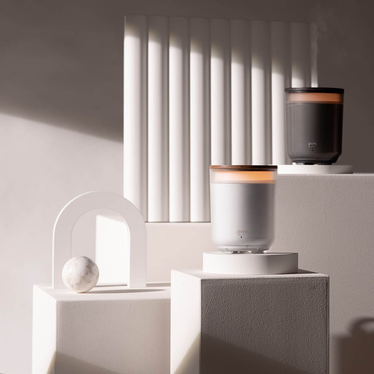 Minimalistische Szene mit geometrischen weißen Plattformen, einem Marmorbogen und einer Kugel, dazu zwei AIR SONIC ipuro Elektrischer Aroma-Diffusor von ipuro - einer weiß, einer schwarz -, der Dampf gegen vertikale weiße Platten abgibt. Perfekt für Duftöle oder Duftkerzen.