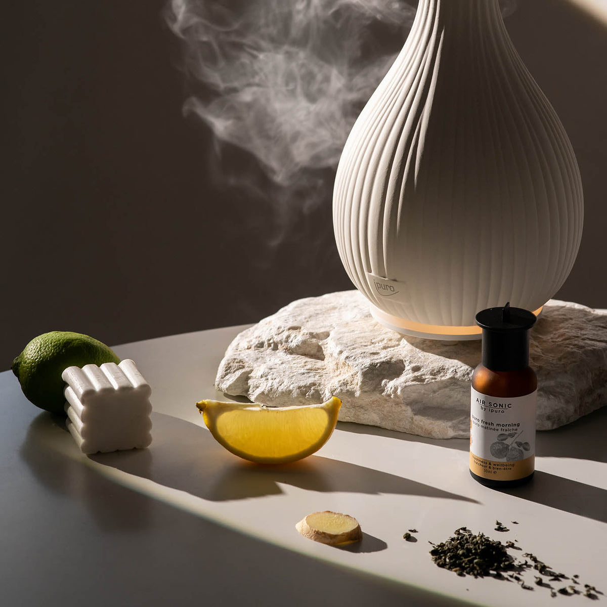 Die AIR SONIC ipuro Aroma-Vase Elektrischer Aroma-Diffusor von ipuro dünstet neben Zitronen- und Limettenscheiben, Ingwer, Teeblättern, Duftöle und einem weißen Strukturartikel auf Stein und glatten Flächen in sanftem Licht aus.
