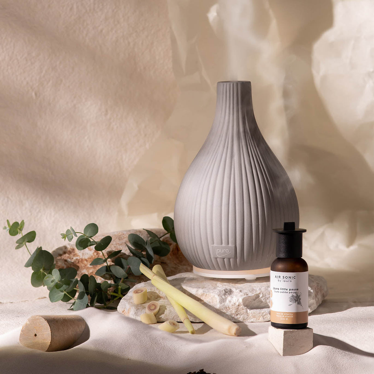 Die AIR SONIC ipuro Aroma-Vase Elektrischer Aroma-Diffusor von ipuro aus grauer Keramik ruht auf einem Stein mit Eukalyptusblättern, Zitronengrasstängeln und einem Duftöle-Fläschchen in der Nähe vor einem weichen beigen Hintergrund.