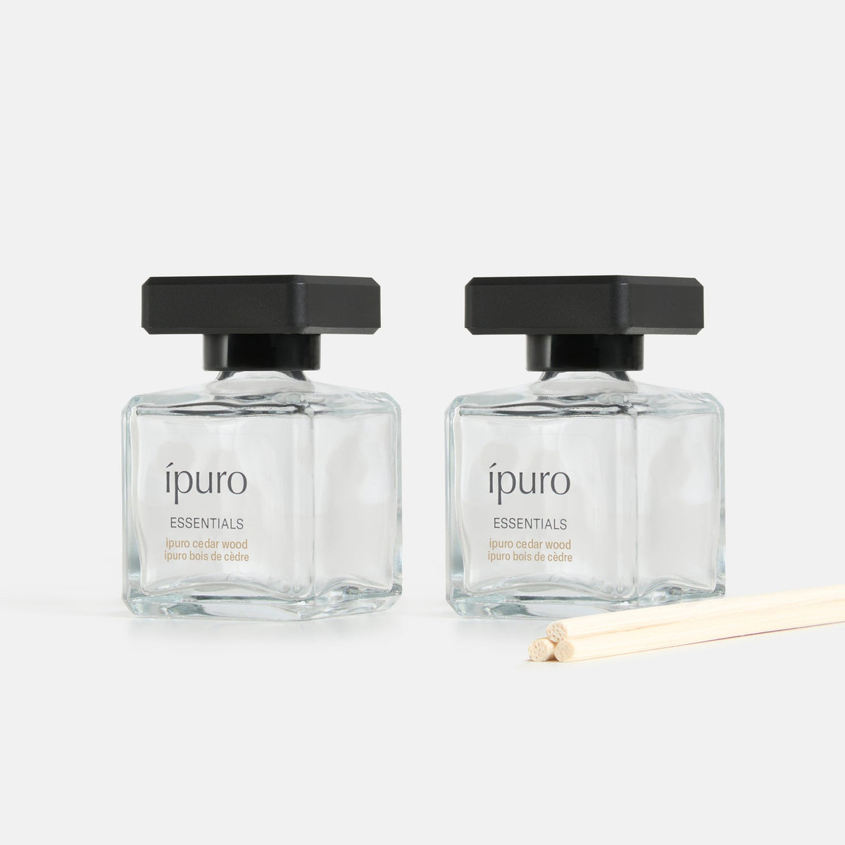 Zwei klare Glasflaschen mit schwarzen Verschlüssen, beschriftet mit "ipuro cedar wood" von ipuro, stehen nebeneinander auf einer weißen Fläche. Drei helle Schilfrohrstäbchen davor verströmen den frischen und beruhigenden Duft von Zedernholz.