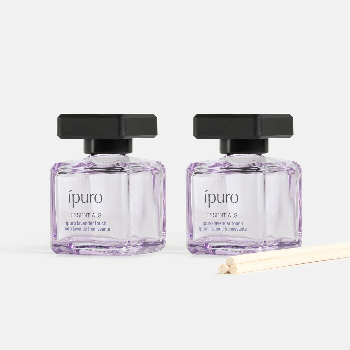 Zwei quadratische Glasflaschen mit schwarzem Deckel, gefüllt mit hellvioletter Flüssigkeit, beschriftet mit ipuro Raumduft-Set lavender touch by ipuro, und davor ein Bündel Schilfrohr-Diffusorstäbchen, ausgestellt vor einem weißen Hintergrund.