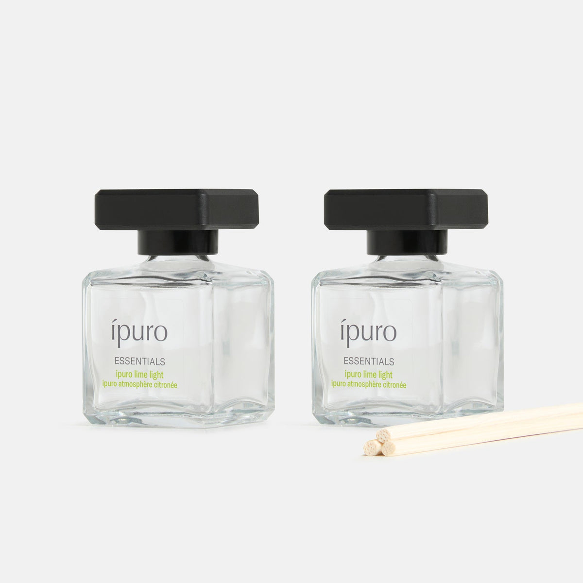 Zwei quadratische ipuro-Glasflaschen aus dem Raumduft-Set ipuro lime light, jeweils mit schwarzem Deckel, stehen neben unbenutzten Diffusorstäbchen auf einer weißen Fläche - ideal, um den Raum mit erfrischender Wellness zu erfüllen.