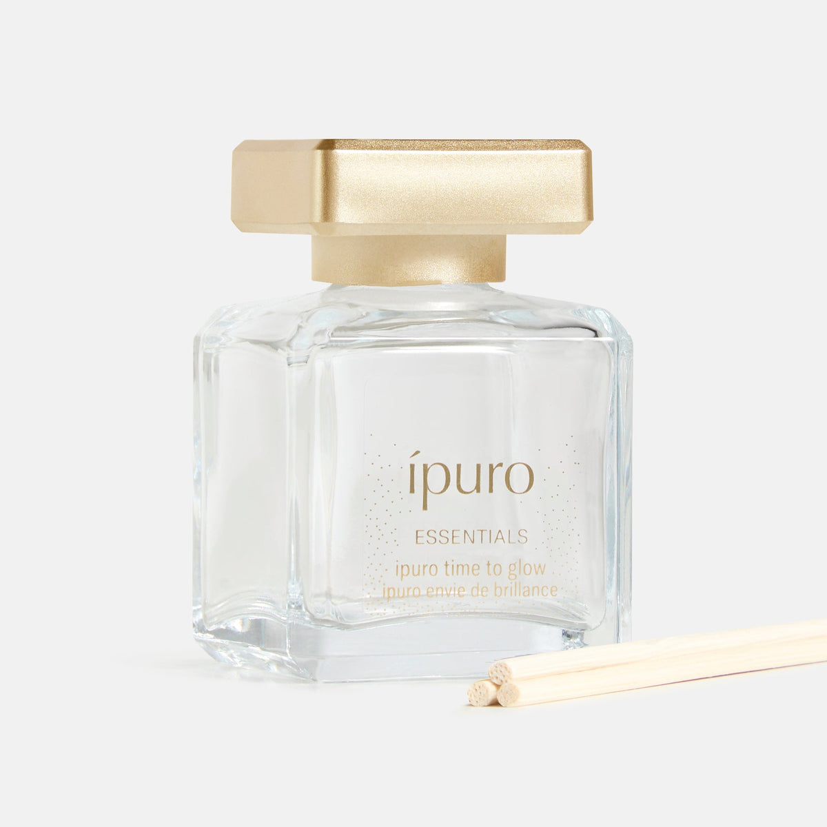 Ein klarer Glasflakon von ipuro Raumduft time to glow, mit goldener Kappe und Diffusorstäbchen, steht auf weißem Hintergrund und erinnert an orientalisch-blumige Noten und die Wärme der Tonkabohne.