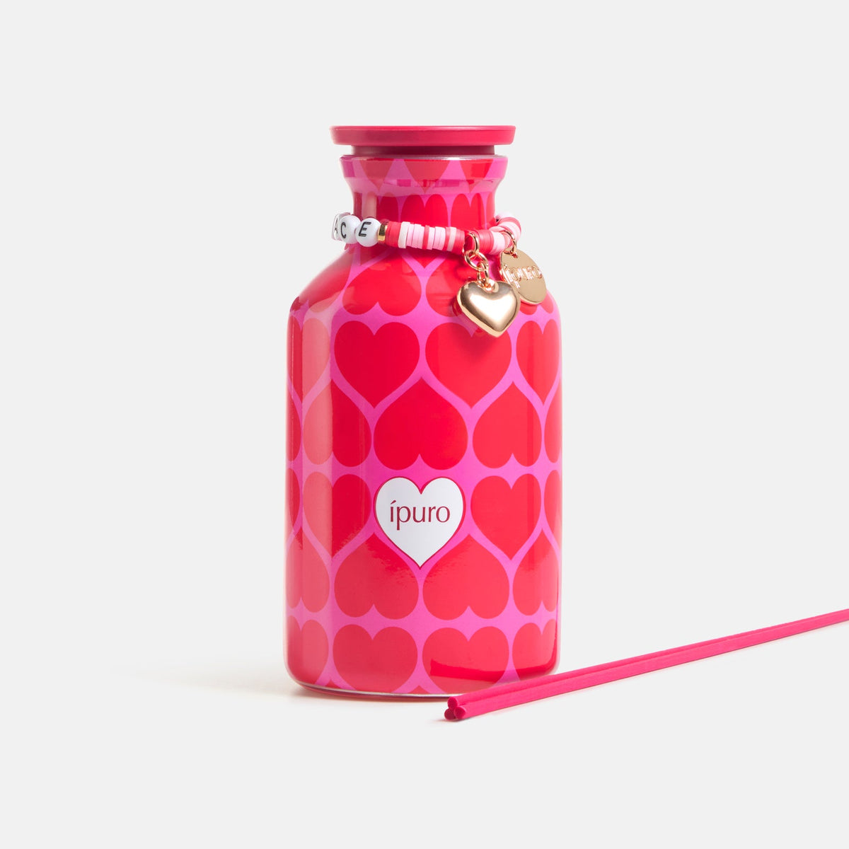 Der ipuro Raumduft love's embrace 240ml hat einen rosafarbenen Flakon mit roten und rosafarbenen Herzen, ein weißes ipuro-Etikett, ein goldenes Herzarmband am Hals und einen passenden Stick - ein bezauberndes Geschenk, inspiriert von seinem zarten Blumenbouquet-Duft.